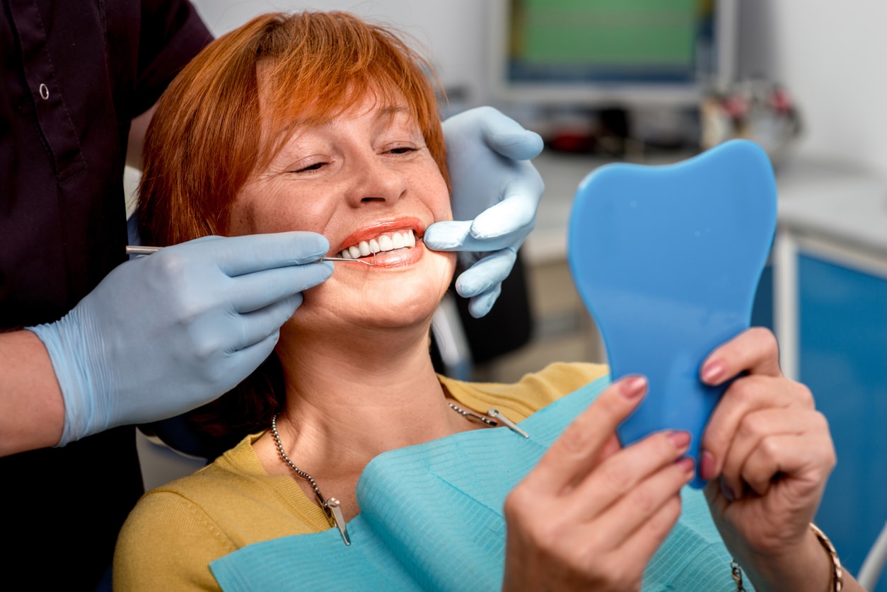 A red-haired woman examining her same-day dentures in a dentist's office chair, holding a mirror.