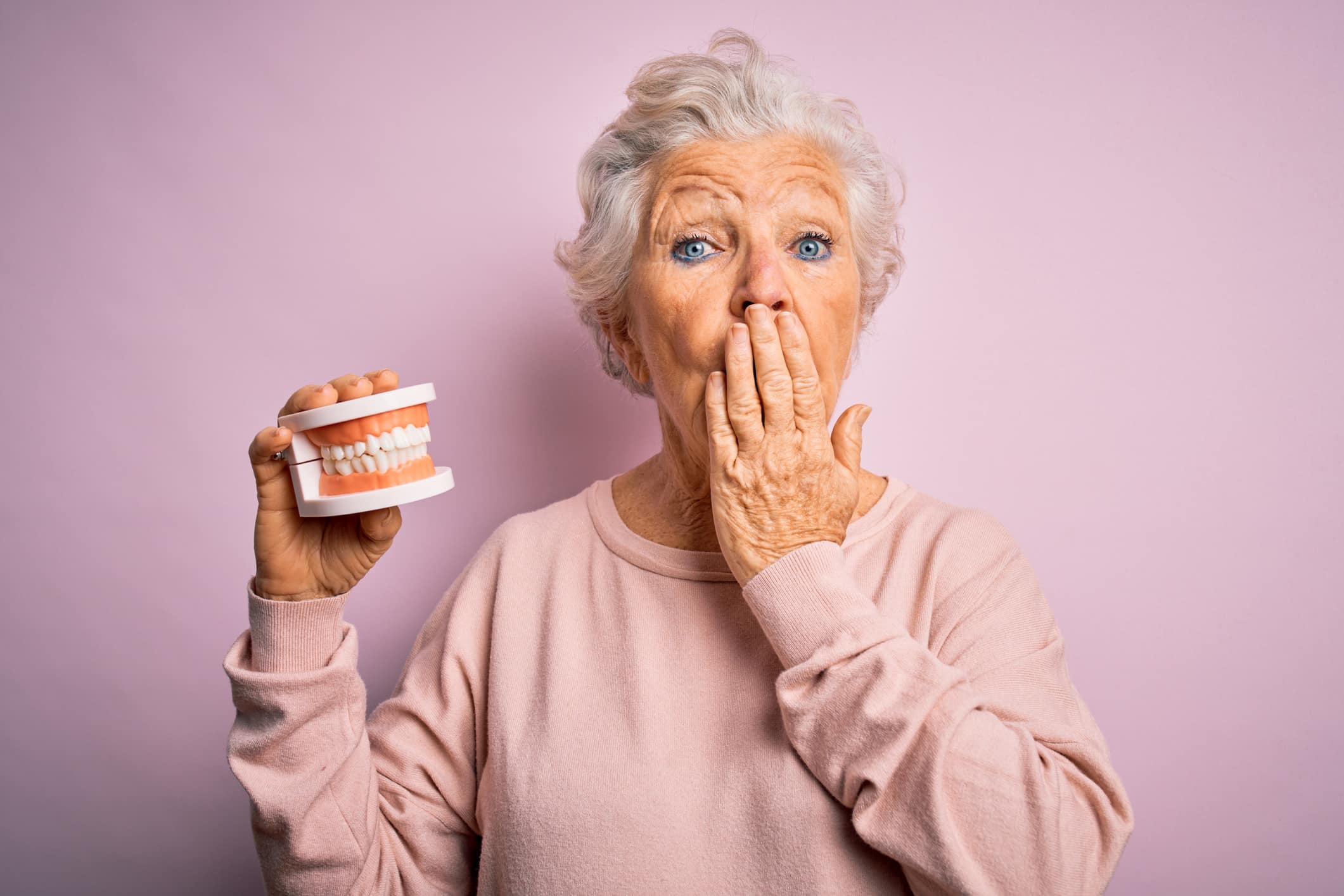 A woman holding a pair of dentures and covering her mouth in front of a pink background.