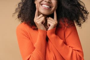 A girl with an orange sweater on, pointing to her healthy smile.