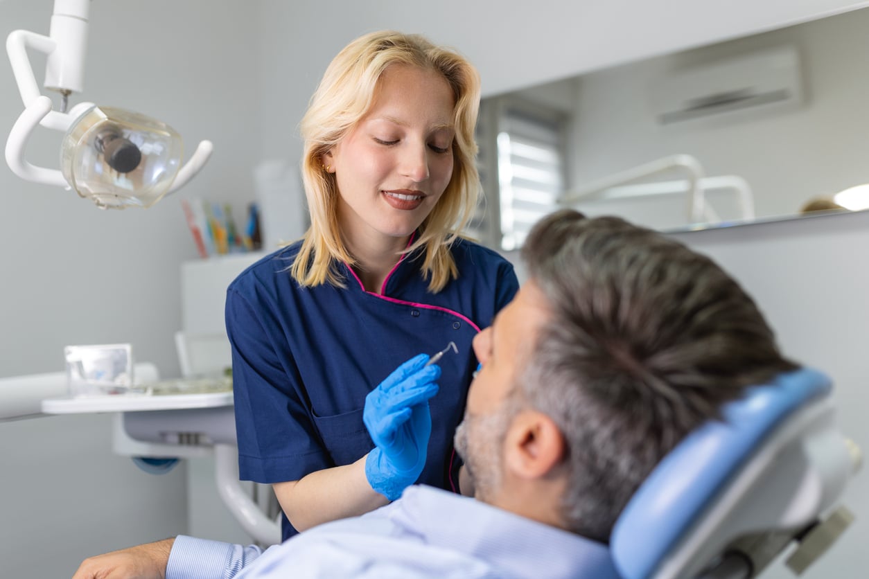 A blonde dentist in blue scrubs doing dental work on a man with brunette hair.