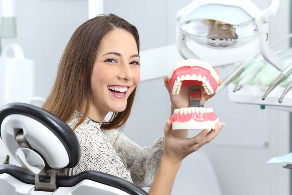 A young woman sitting in a dentist’s chair holding a model of dentures.