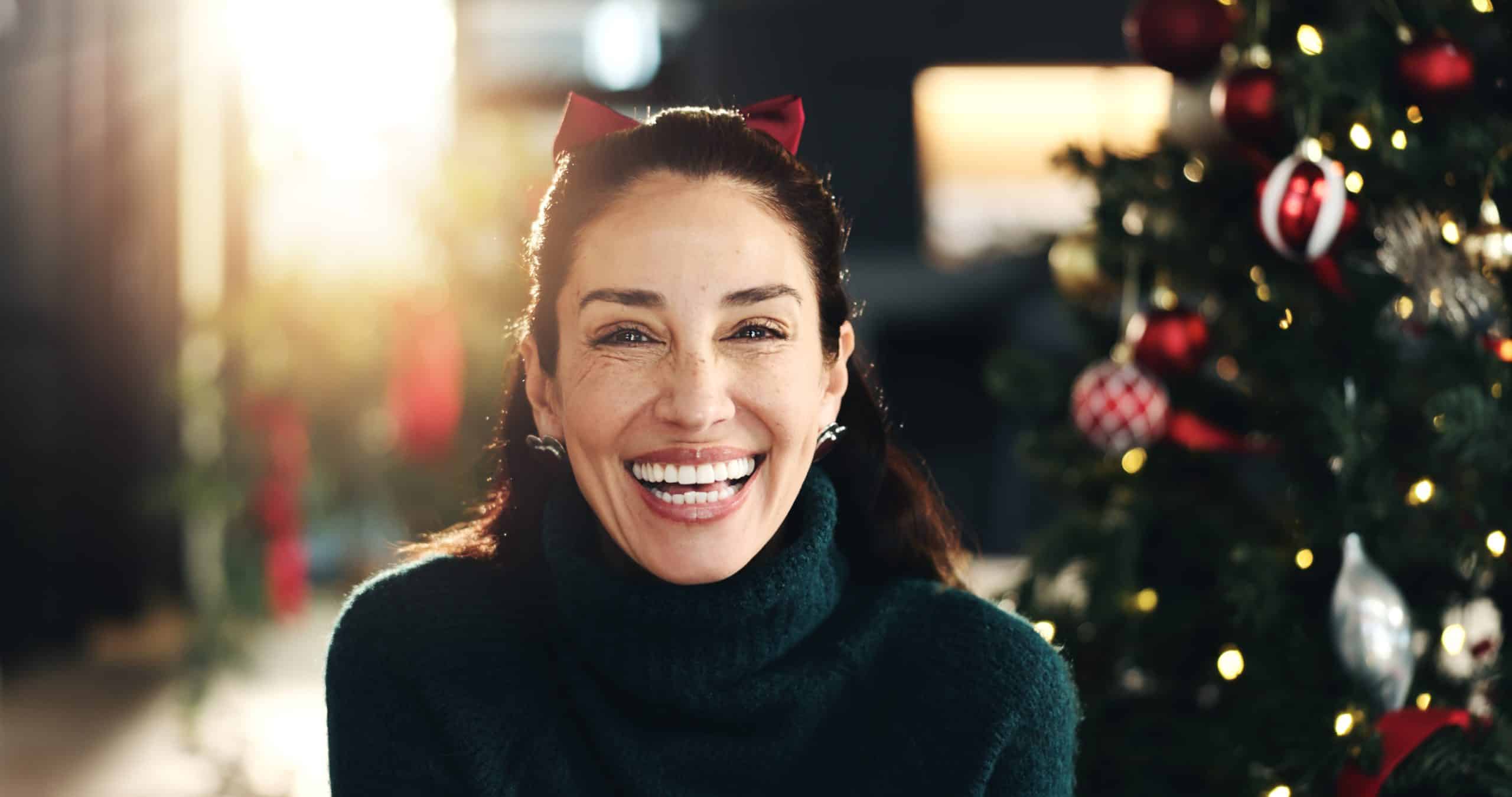 A woman with dentures wears a bright smile in front of a Christmas tree and other holiday decorations.